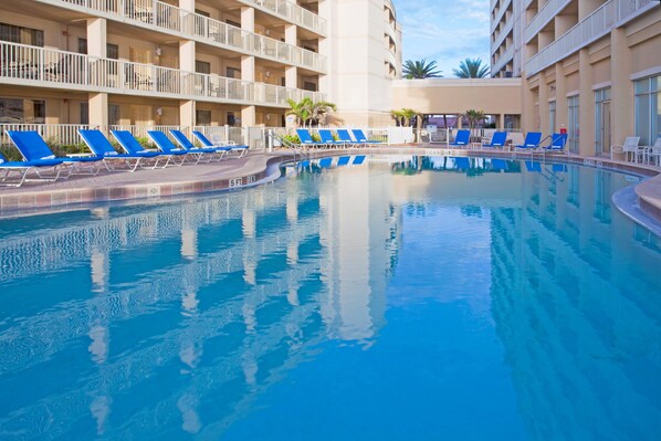 Una piscina con sillas de descanso azules, una cerca blanca y un edificio de varios pisos con balcones.