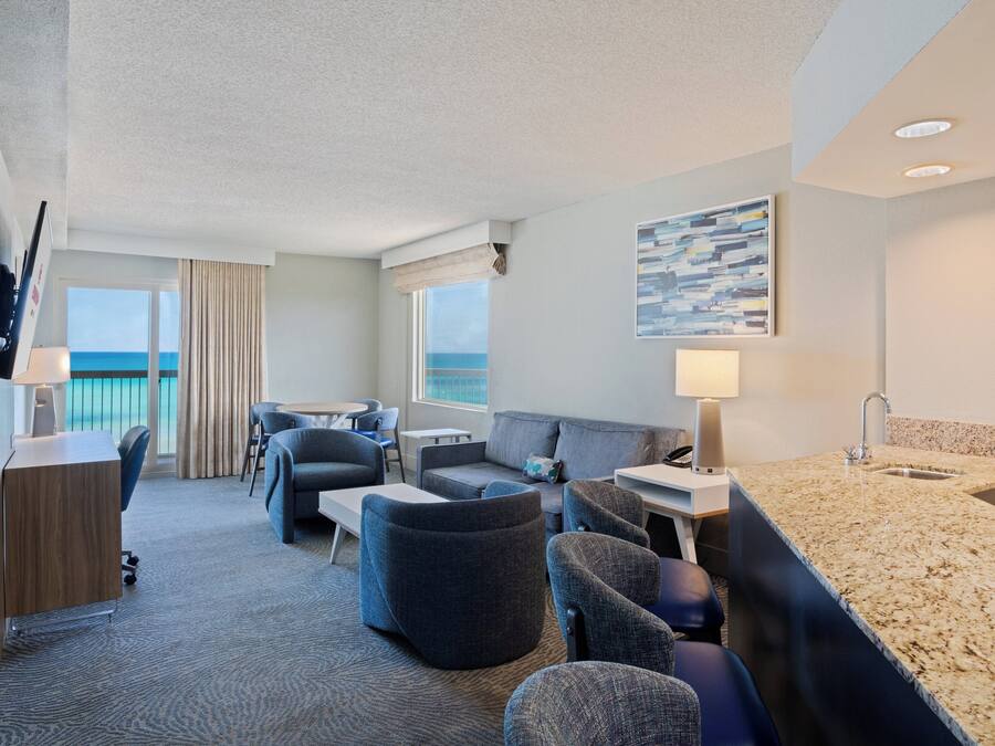 A hotel room with a balcony, a sofa, a desk, and a kitchenette.