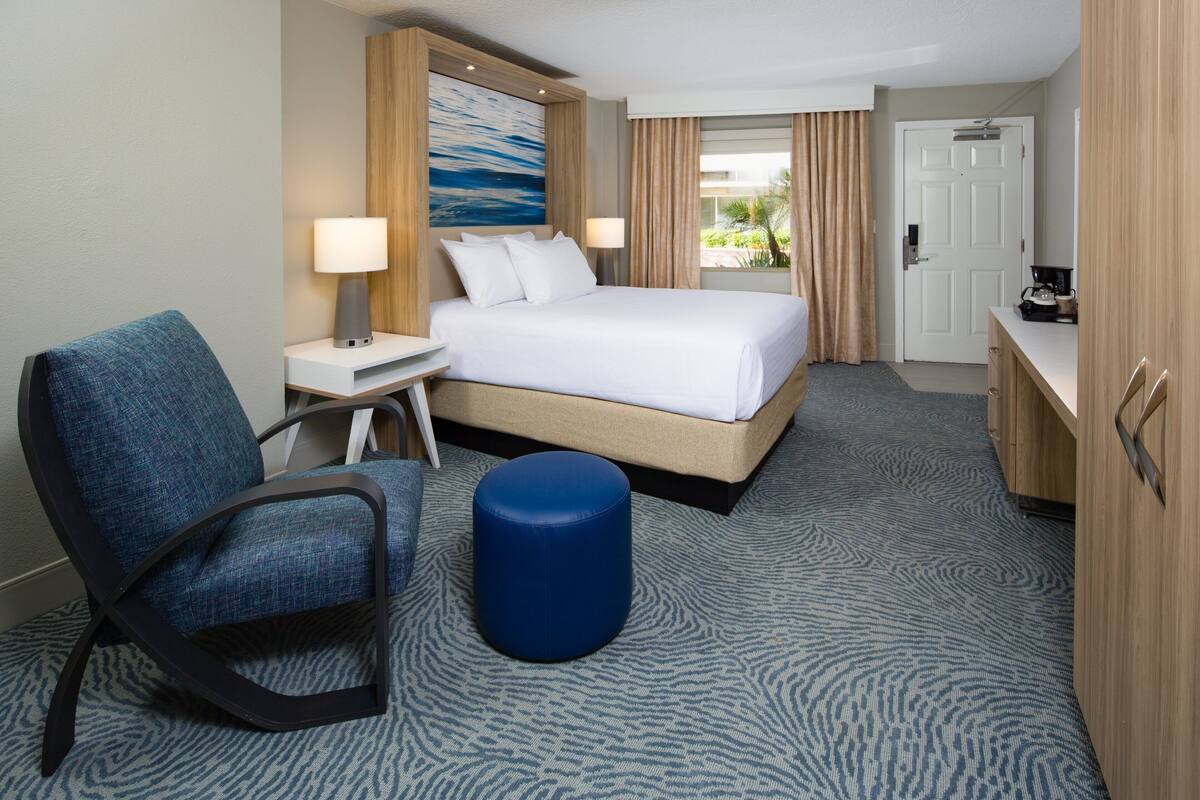 A hotel room with a bed, a desk, a chair, a blue ottoman, and a view of the outdoors.