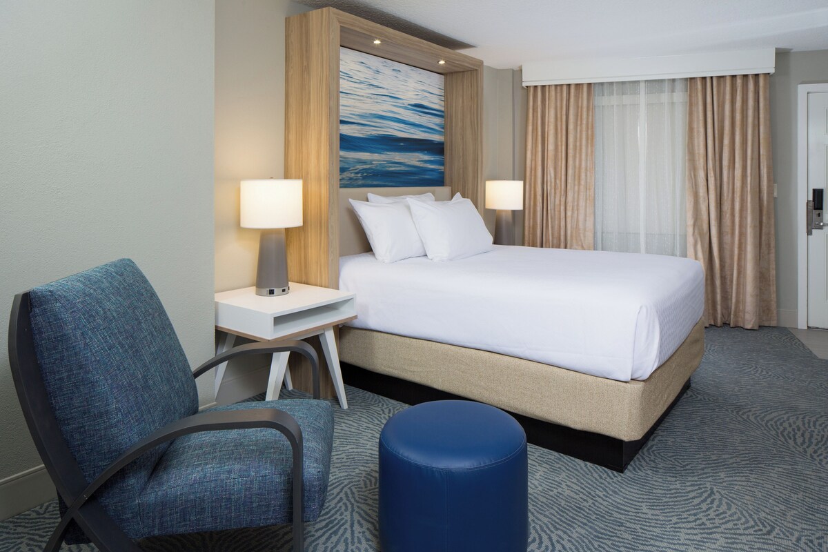 A hotel room with a bed, two bedside lamps, a chair, a desk, a blue ottoman, and a large painting on the wall.