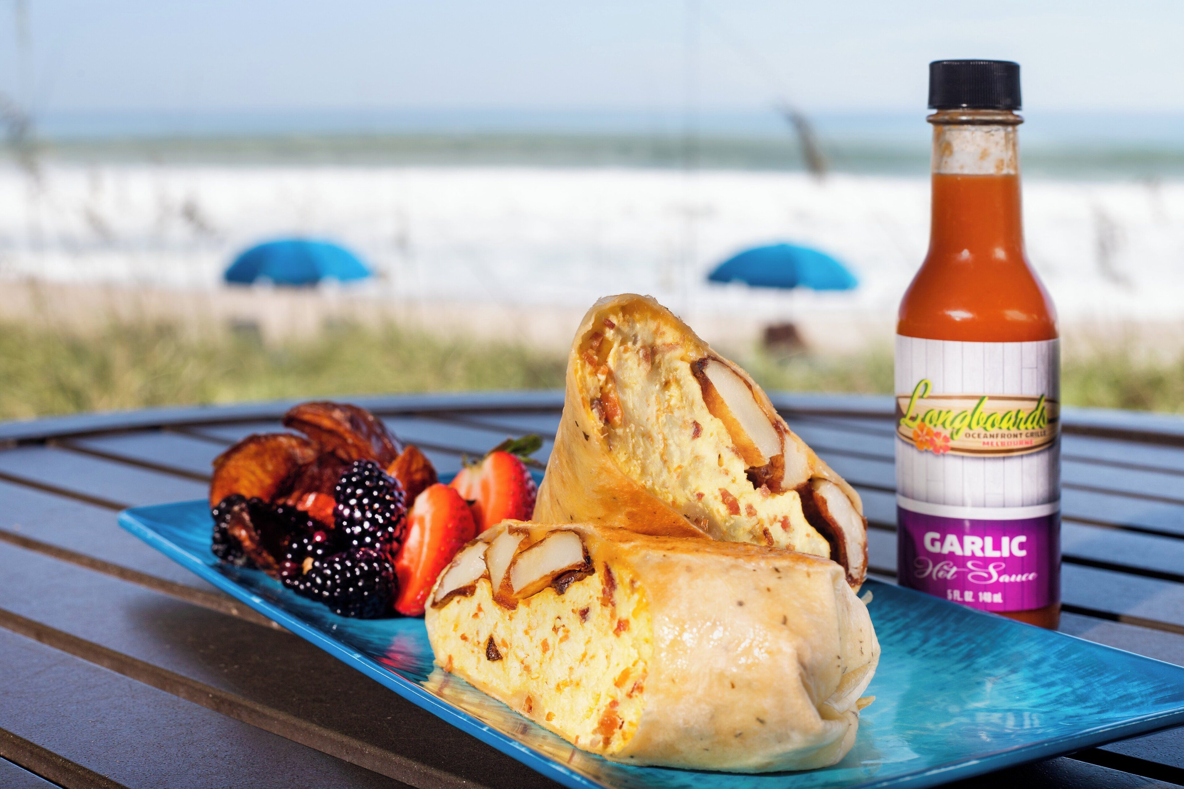 A plate with food and a bottle of sauce on a table with a beach view in the background.