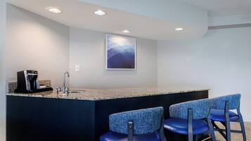 A bar area with a granite countertop, two blue upholstered bar stools, a coffee machine, and a wall-mounted artwork.