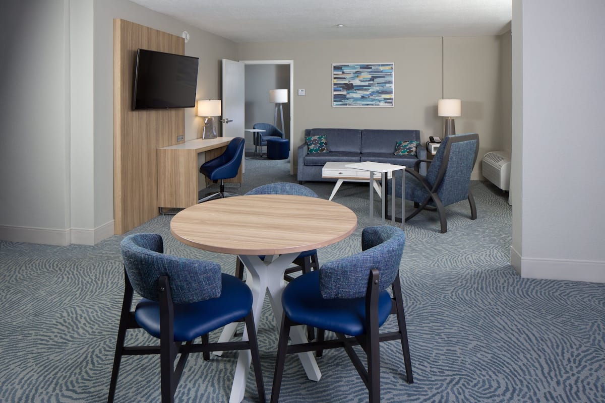 A hotel room with a round table, blue chairs, a sofa, a TV, and a painting on the wall.