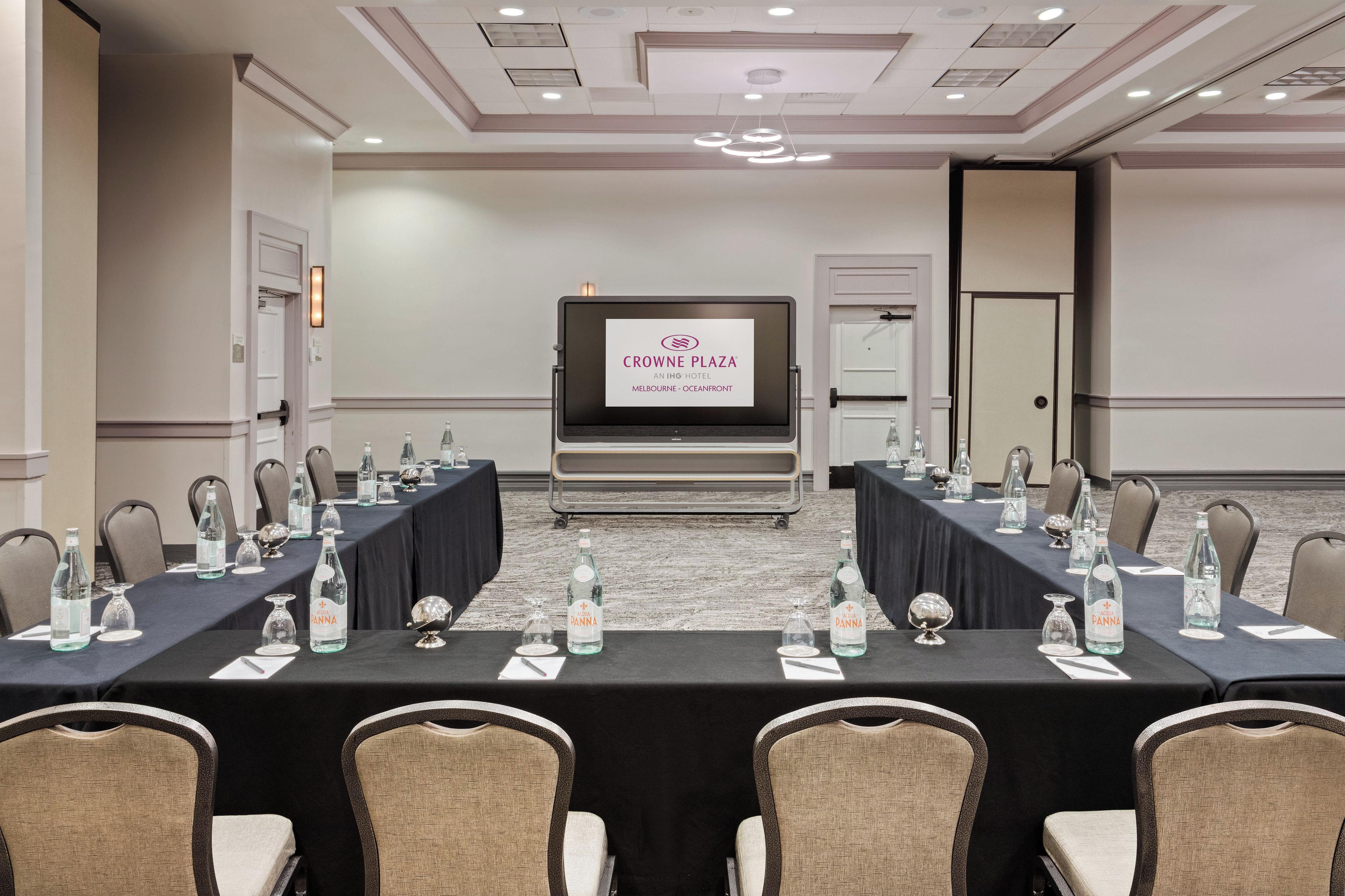 A conference room with a long table, chairs, water bottles, and a screen displaying 'Crowne Plaza.'