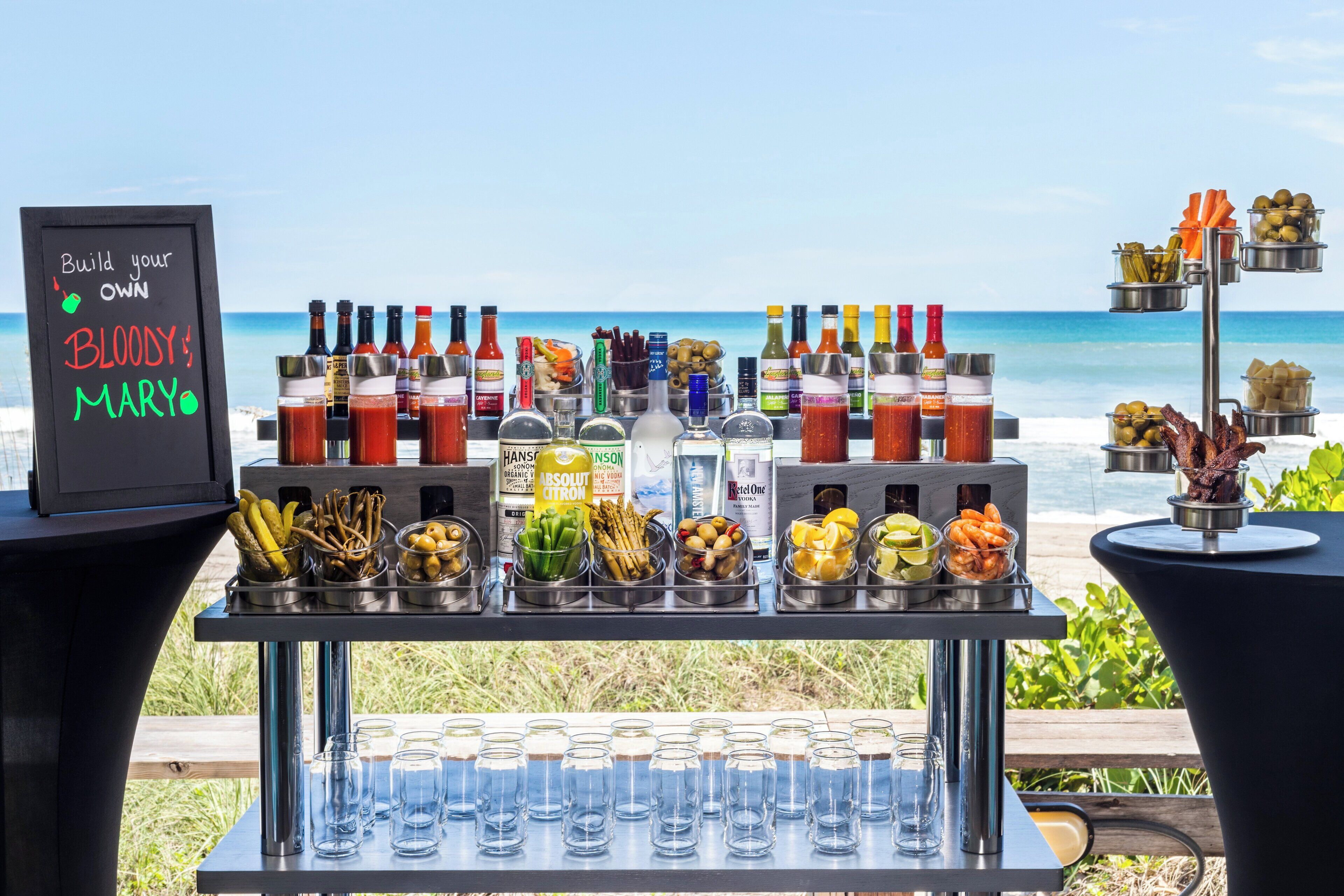 A bar setup with various condiments, bottles, and glasses, with a view of the ocean in the background.