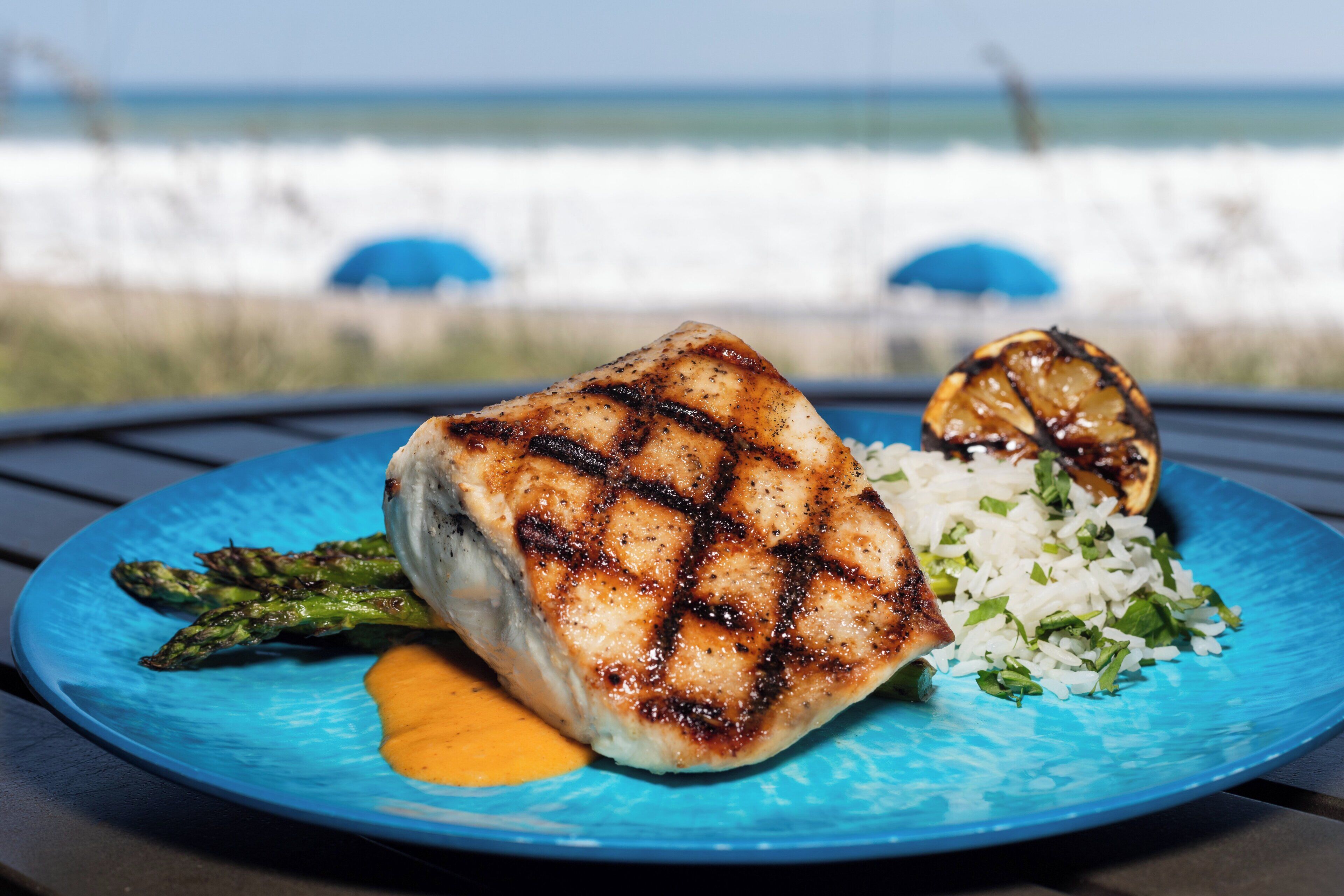 Ein Teller mit gegrilltem Hähnchen, Spargel und Reis, im Hintergrund ist der Strand zu sehen.