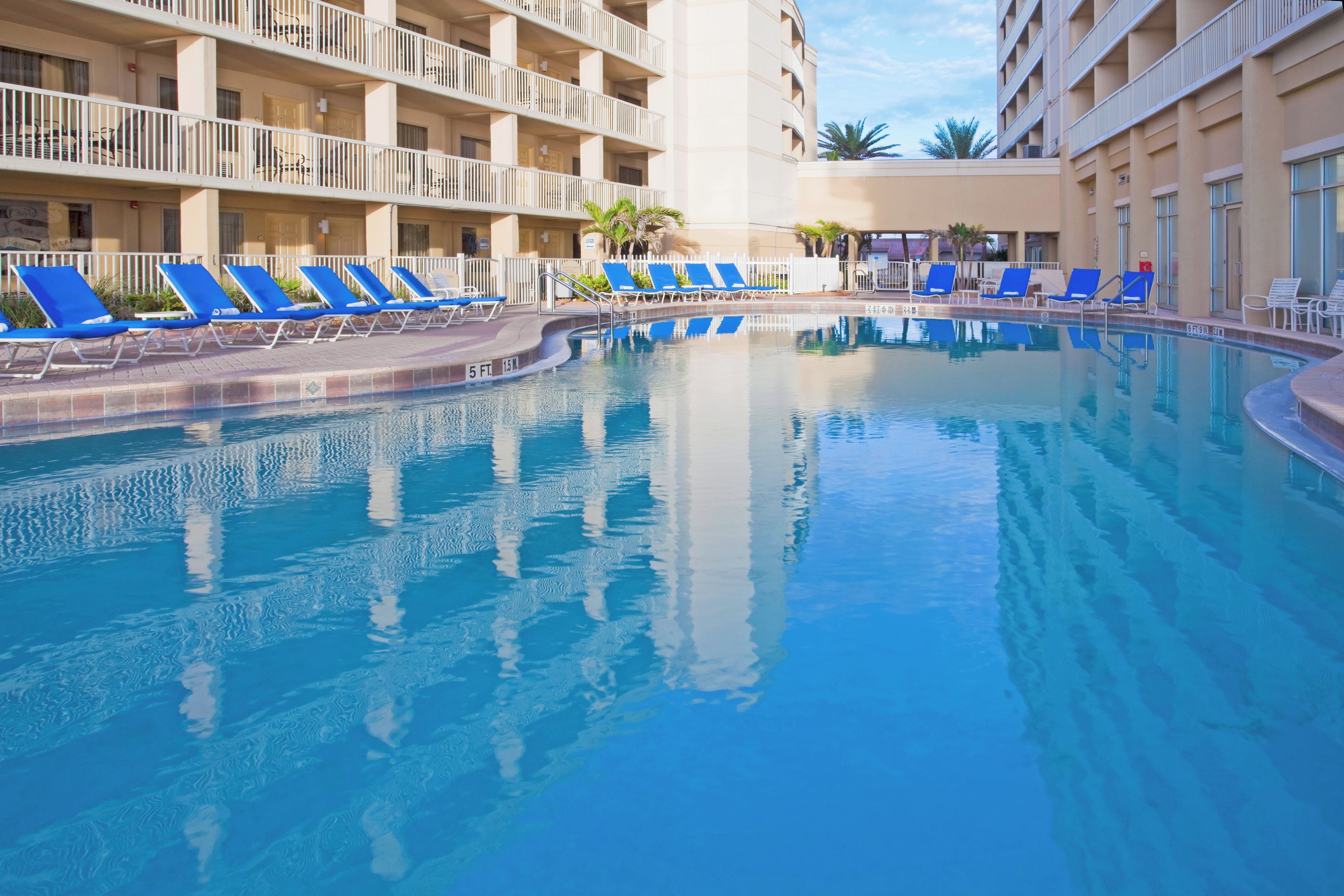 A swimming pool with blue lounge chairs, a white fence, and a building with balconies in the background.