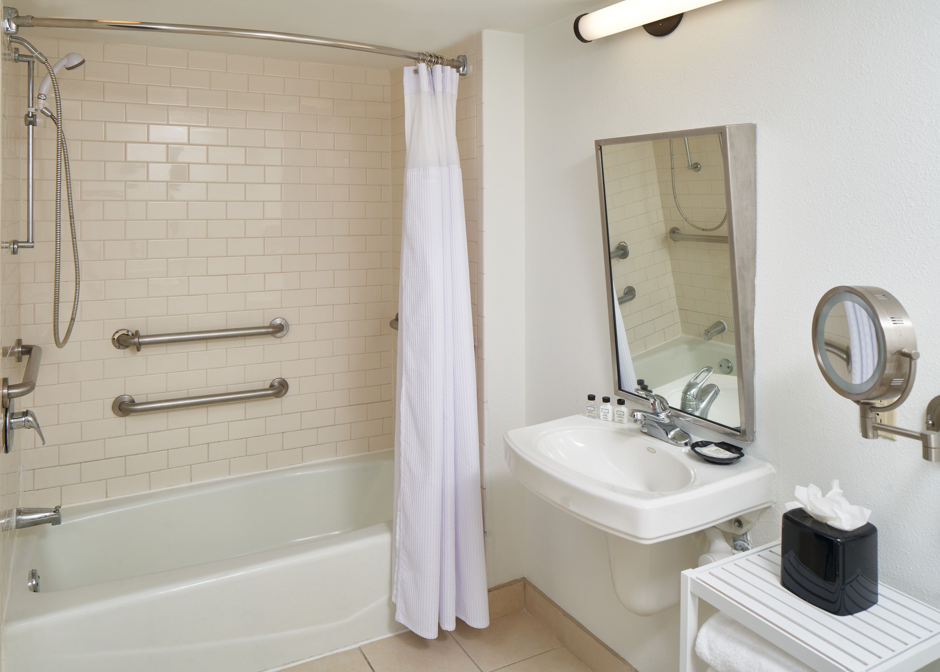 A bathroom with a bathtub, shower, white tiled walls, a sink with a mirror, and a towel dispenser.