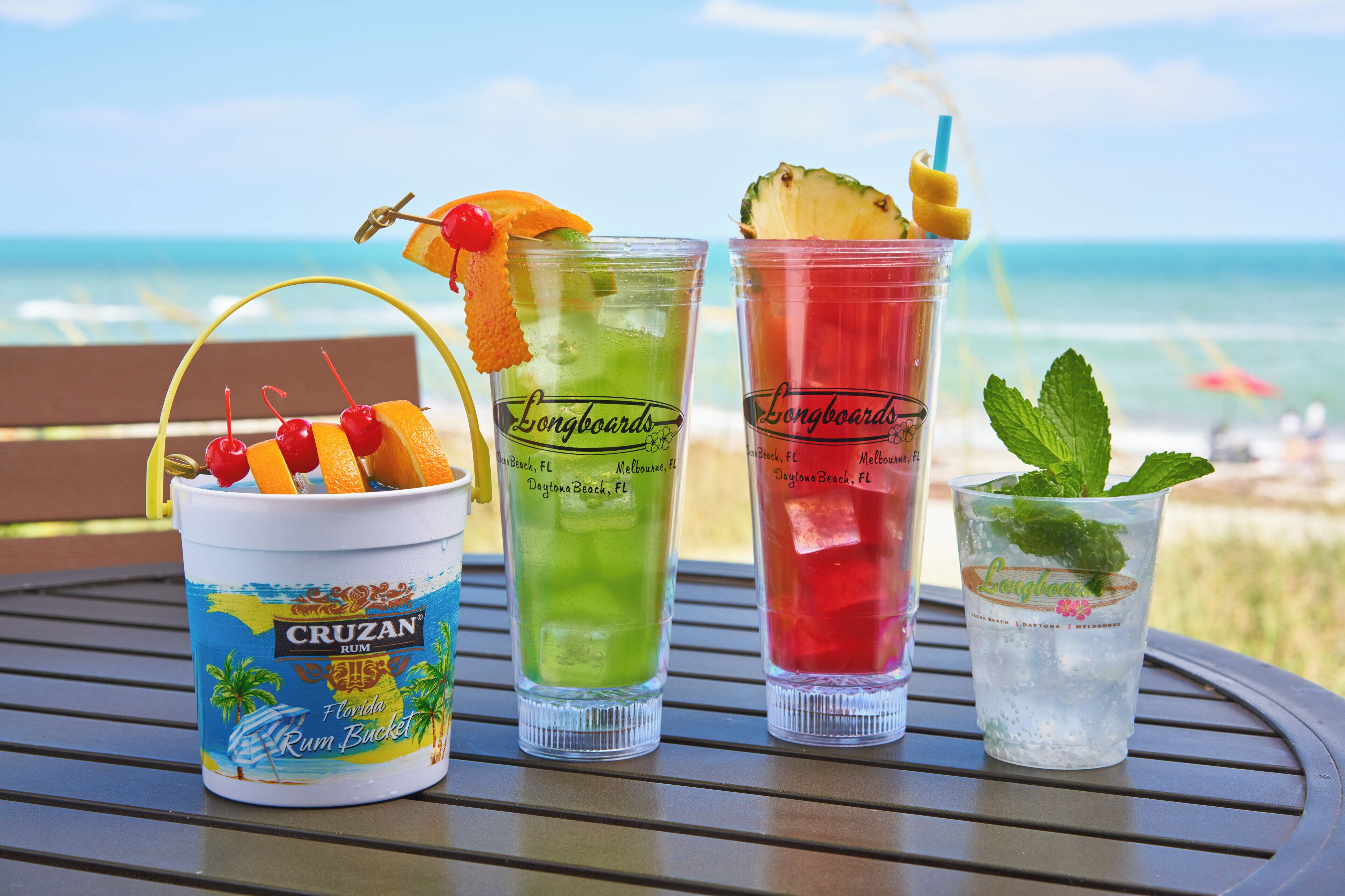 Trois récipients contenant des boissons sont posés sur une table, avec la plage et l’océan en arrière-plan.