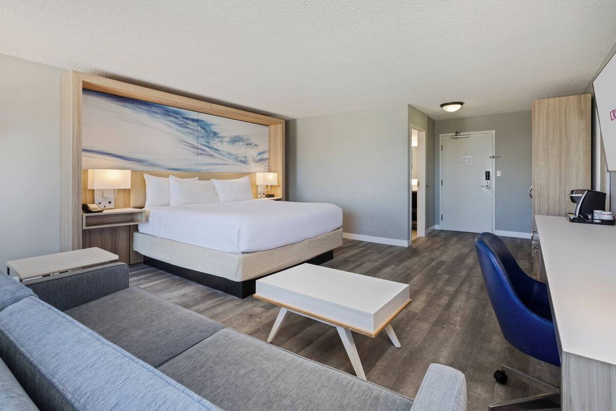 A modern hotel room with a large bed, a desk, a sofa, and a painting on the wall.