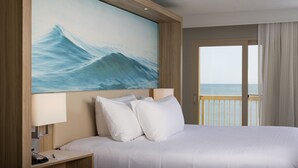 Camera d'albergo con un letto grande, un comodino con una lampada, un quadro raffigurante un'onda e vista sul mare da un balcone.