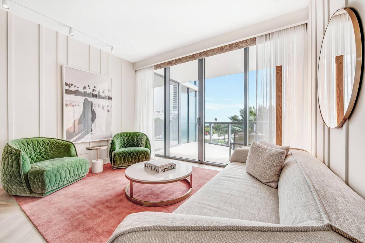 A modern living room with a green sofa, a beige sofa, and a round coffee table. There is a large window with a view of buildings and a balcony.