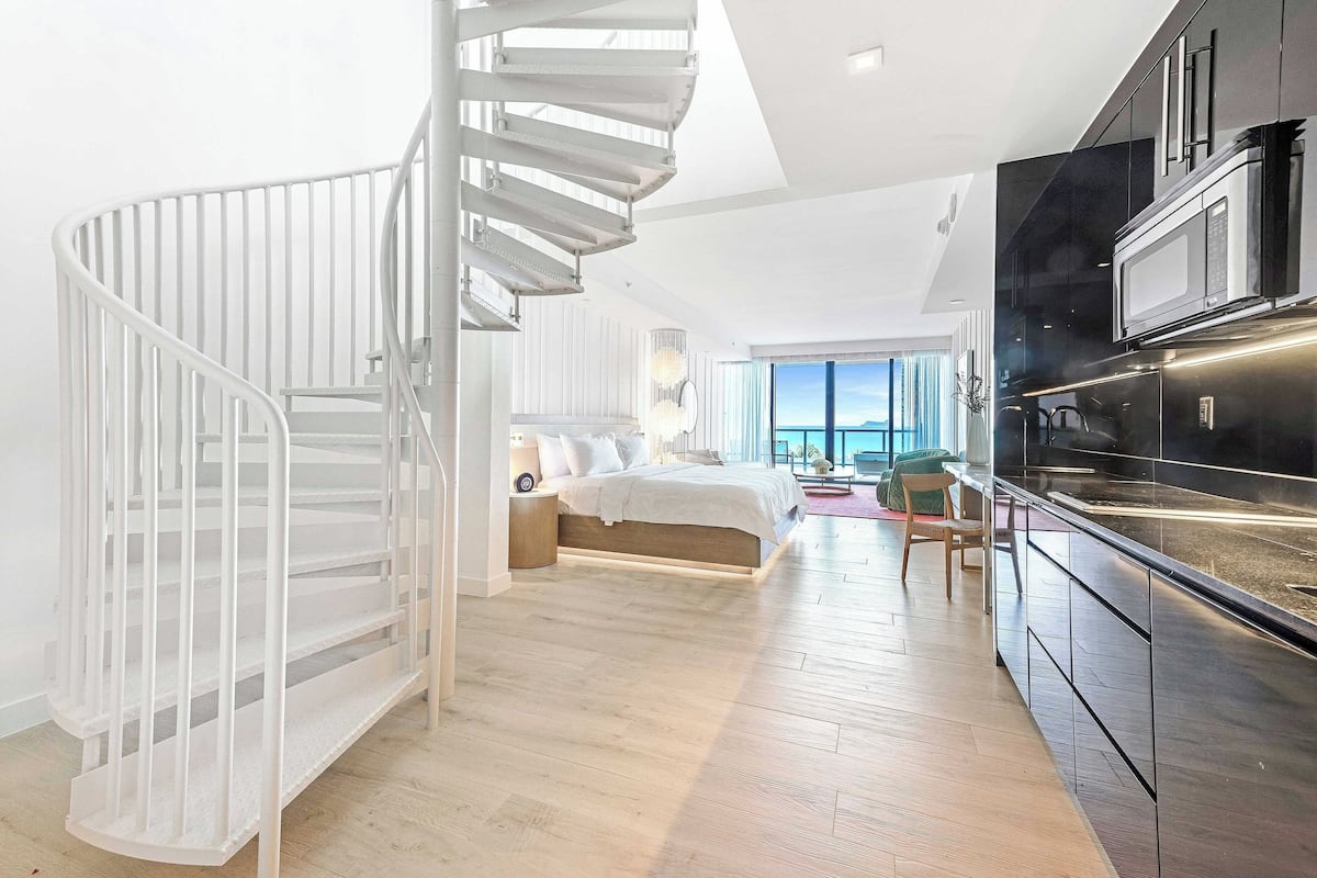 A modern interior with a spiral staircase, a large bed, a kitchen with dark cabinetry, and a view through the window.