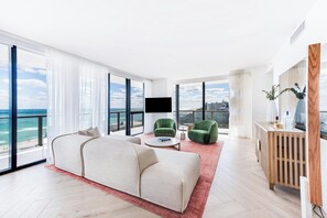 A spacious living room with a large sofa, two green armchairs, a coffee table, and a TV. The room has large windows offering a view of the ocean and a cityscape.