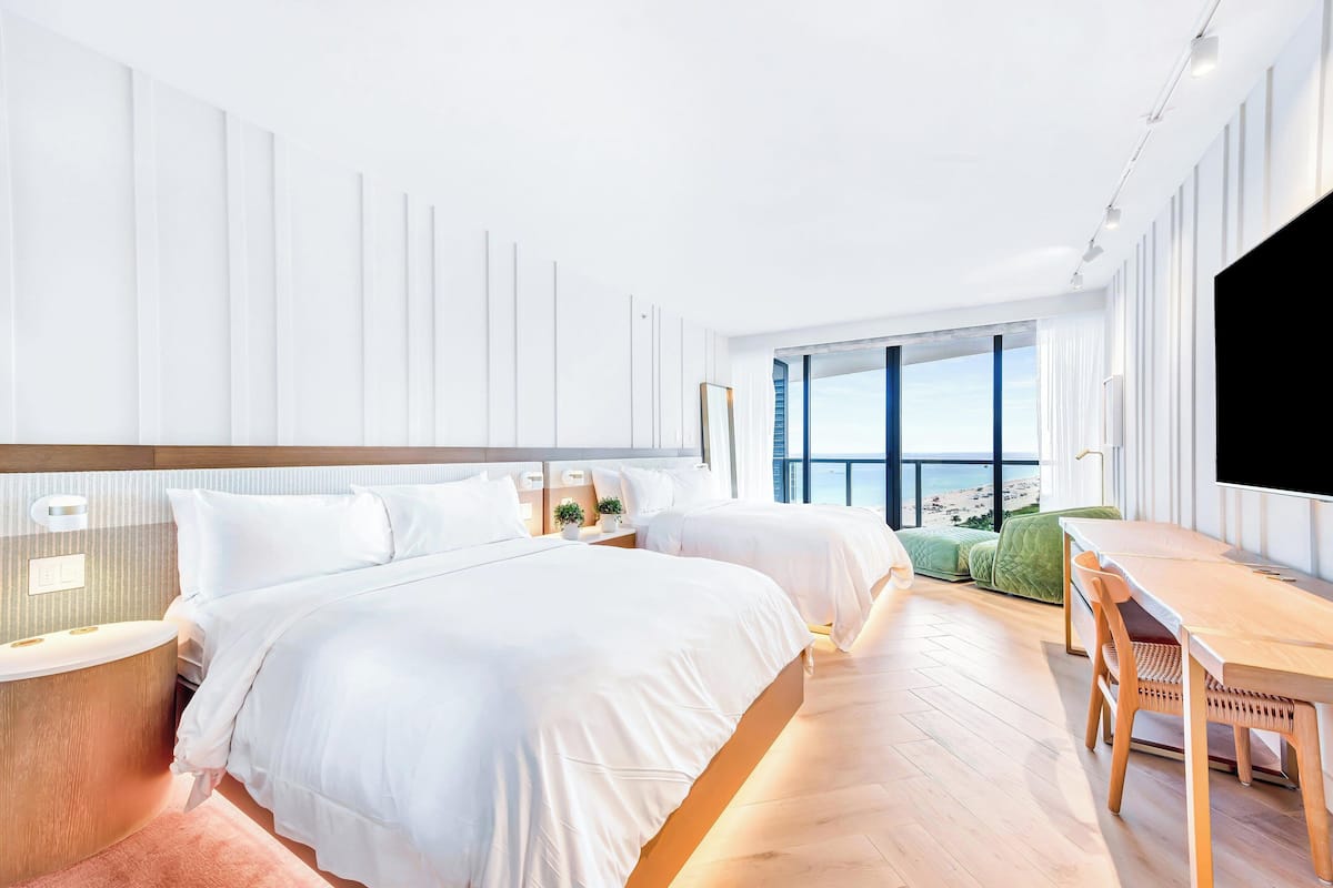 A modern hotel room with two beds, a wooden dining table, and a large window offering a view of the beach.