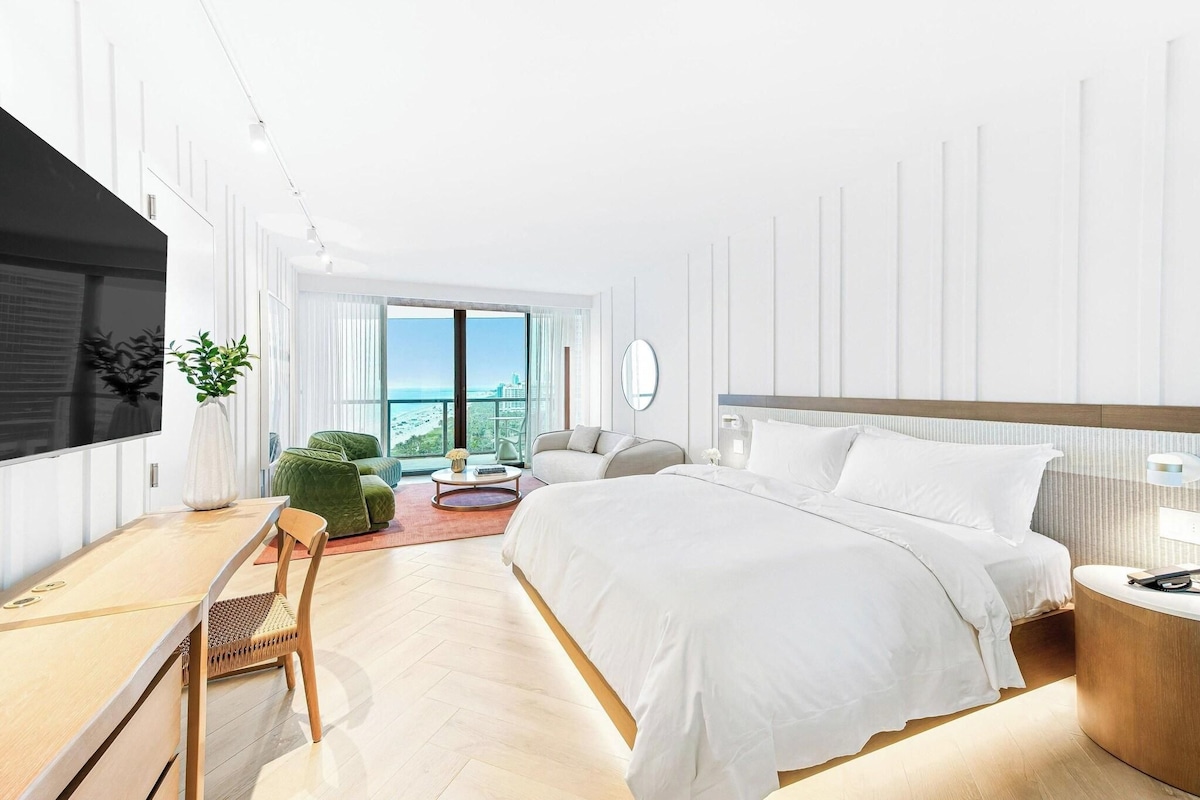 A modern hotel room with a large bed, a desk, a chair, a sofa, and a view of the city.