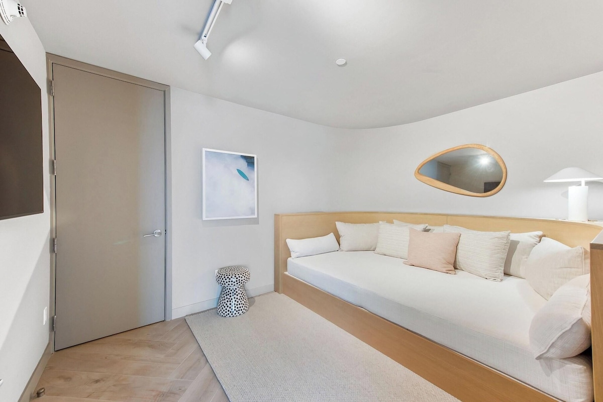 A modern hotel room with a bed, a bedside table, a wall-mounted mirror, and a small round table.
