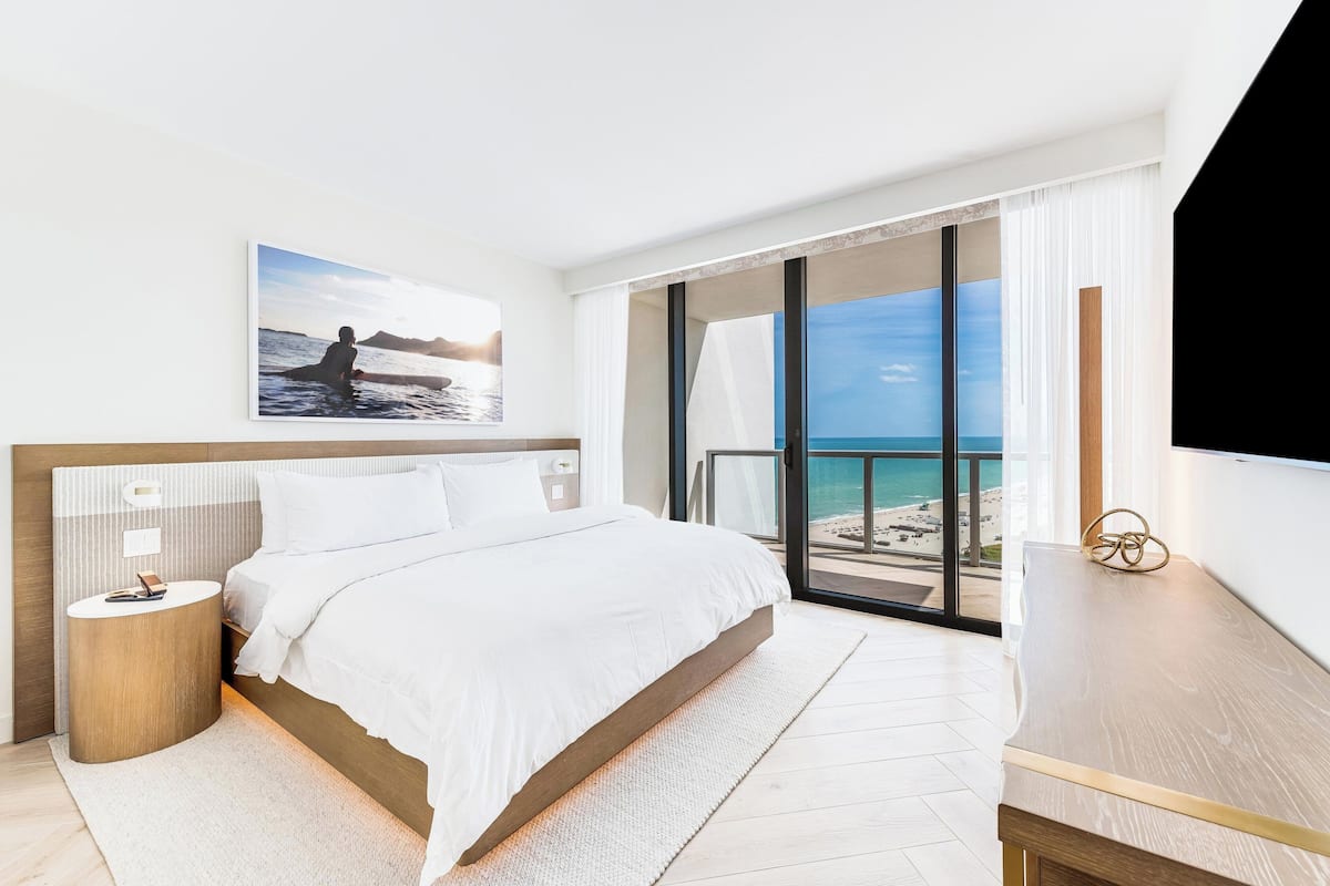 A modern hotel room with a large bed, a bedside table, a nightstand, a flat-screen TV, and a balcony with a view of the beach.