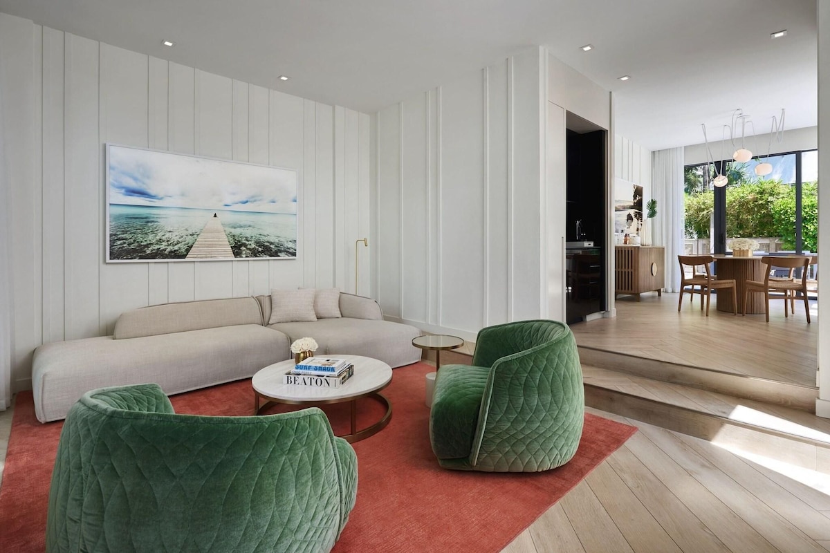 A modern living room with a sofa, green chairs, a coffee table, and a large painting of a pier.