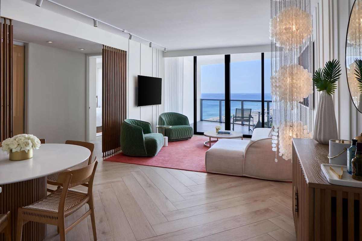 A modern hotel room with a balcony, a round dining table, green chairs, a sofa, and a TV mounted on the wall.