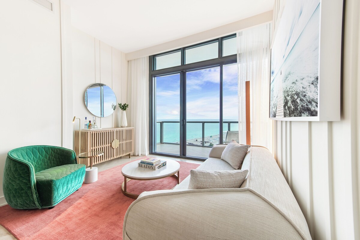 A modern living room with a view of the ocean, a green armchair, a beige sofa, and a round mirror.