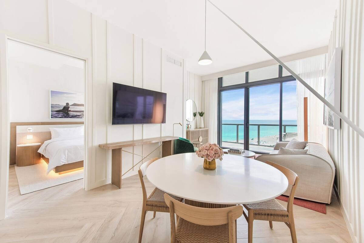 A modern hotel room with a large window, a flat-screen TV, a dining area with a round table, and a cozy seating area.
