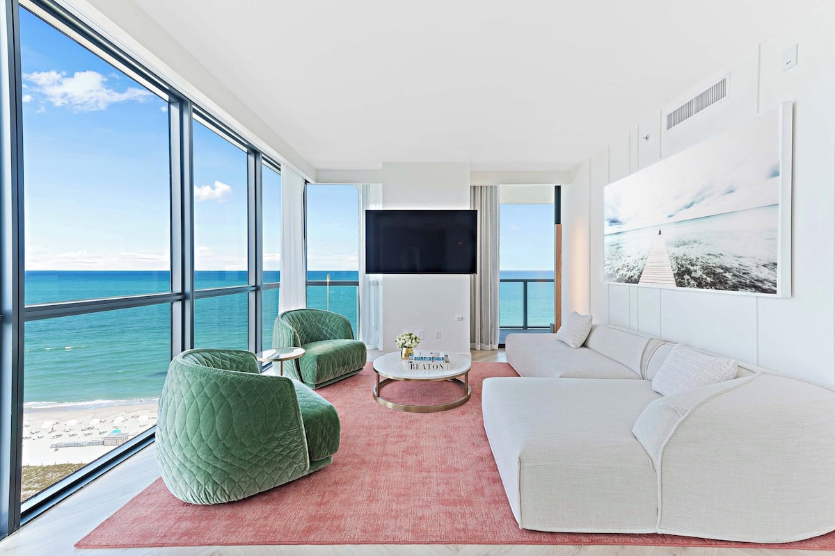 A modern living room with large windows, a white sofa, green armchairs, and a flat-screen TV.