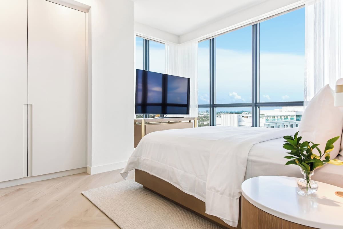 A modern hotel room with a large bed, a flat-screen TV, a round side table, and a view of the city through large windows.