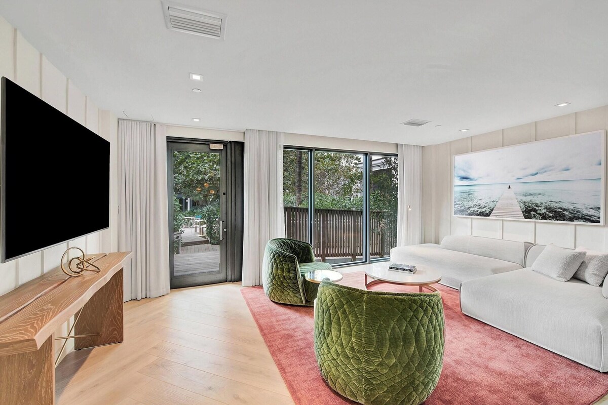 A modern living room with a flat-screen TV, a wooden bench, a green upholstered sofa, a white coffee table, and a large window with a view of greenery.