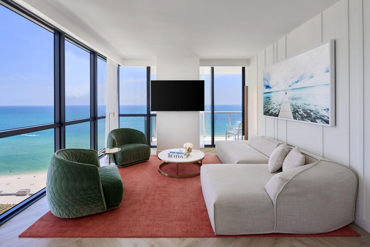 A modern living room with a large window, a flat-screen TV, and a view of the beach.