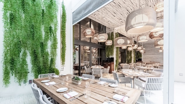 A modern dining area with wooden tables and white chairs, surrounded by hanging greenery and stylish pendant lights. - W South Beach (Miami Beach)