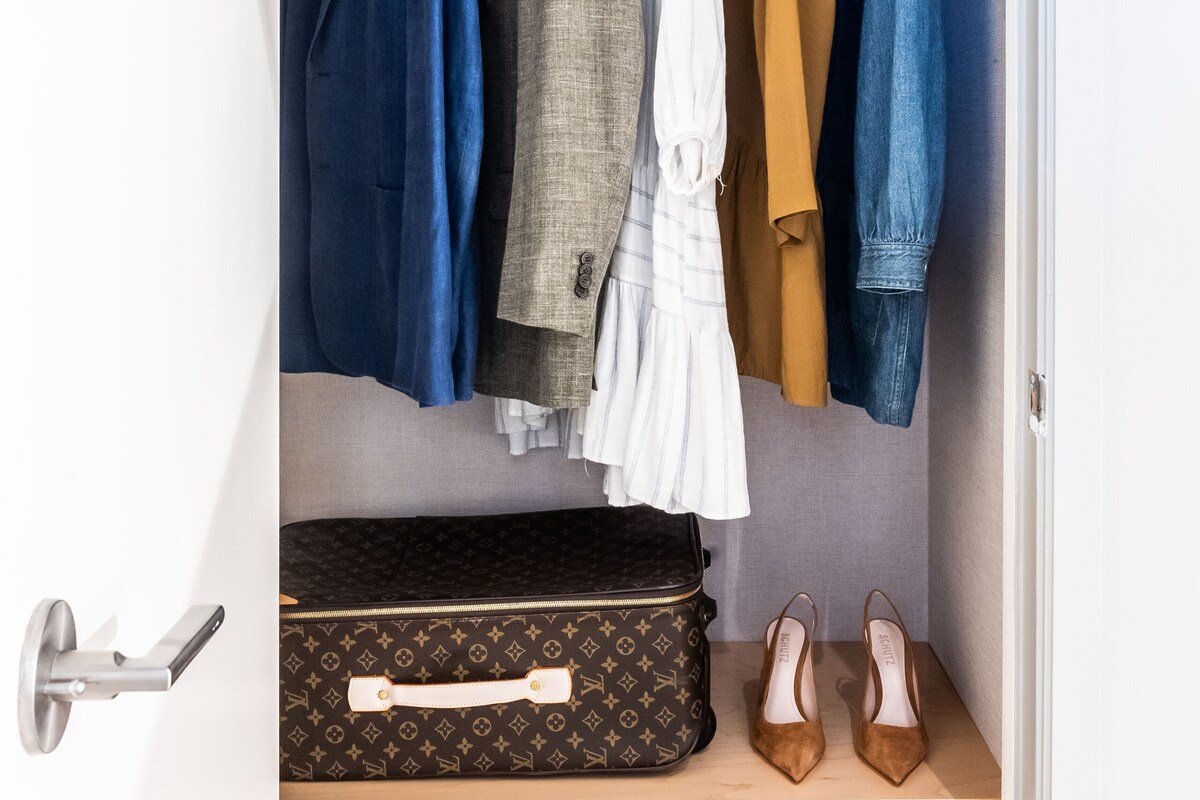 A closet with hanging clothes and a suitcase on a shelf.