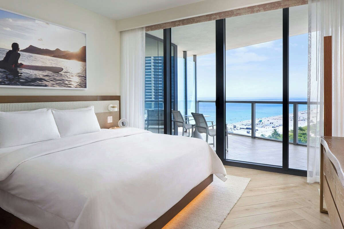 A hotel room with a large bed, a view of the beach, and a balcony with chairs.