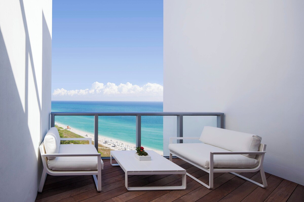 A balcony with a white sofa, a white coffee table, and a view of the beach and ocean.