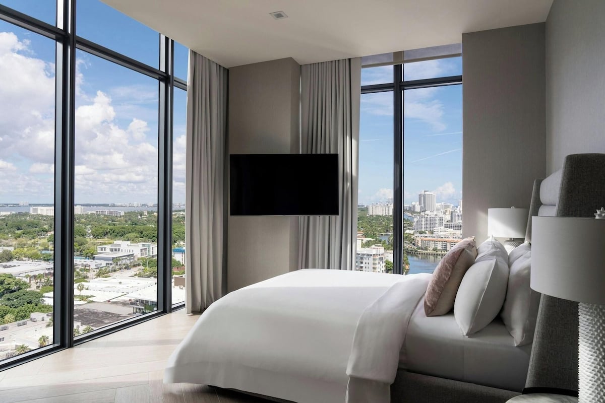 A hotel room with a large bed, a flat-screen TV mounted on the wall, and a view of the cityscape through large windows.