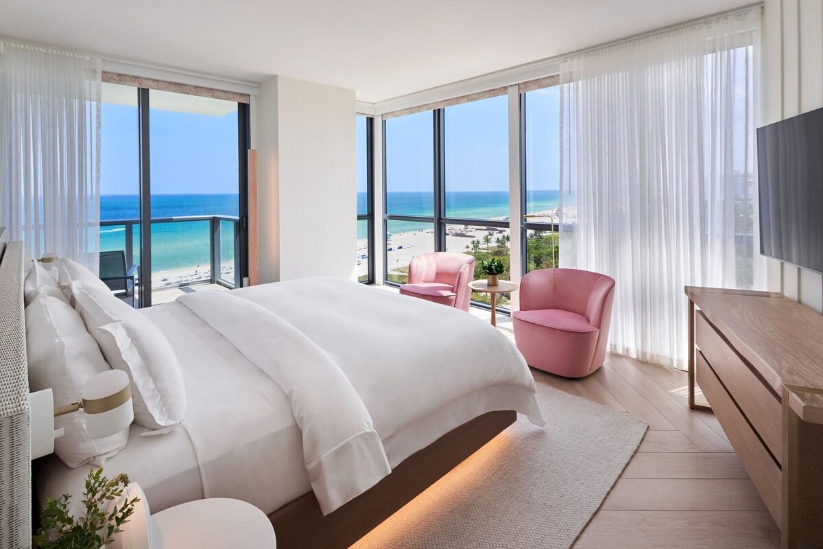 Une chambre d’hôtel moderne avec un grand lit, un fauteuil rose, une télévision et un balcon donnant sur la plage.