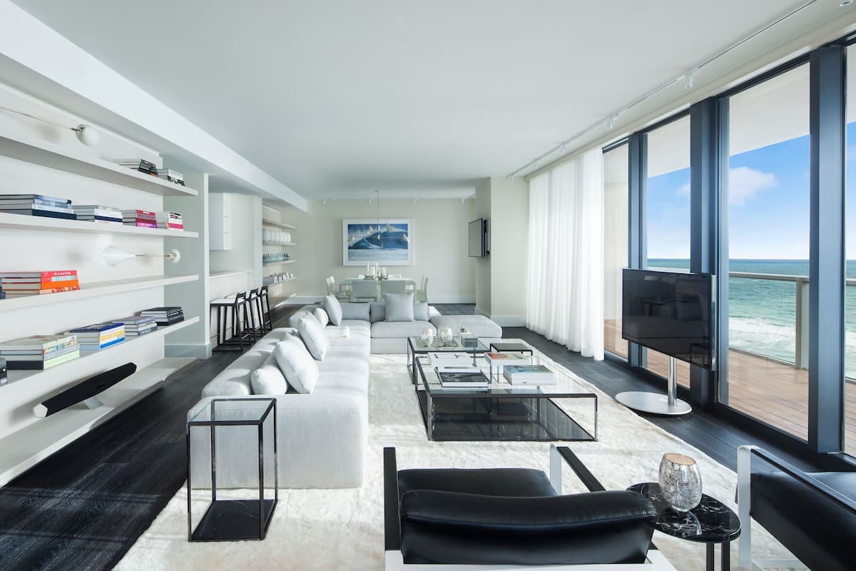 A modern living room with a sofa, coffee table, and TV, large windows with a view of the ocean, and bookshelves.