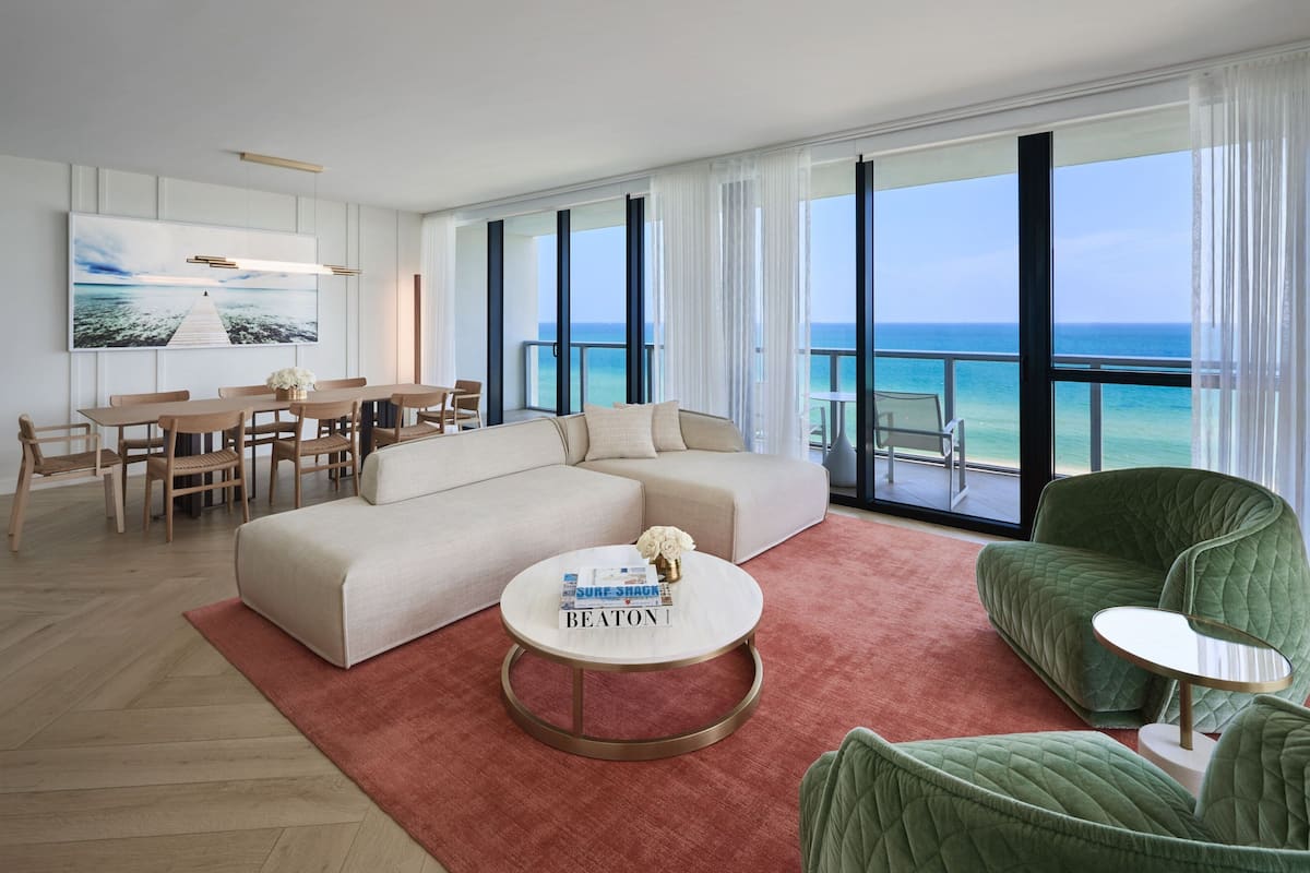 A modern living room with a large sofa, a coffee table, and a dining area with chairs. There is a large window with a view of the ocean.