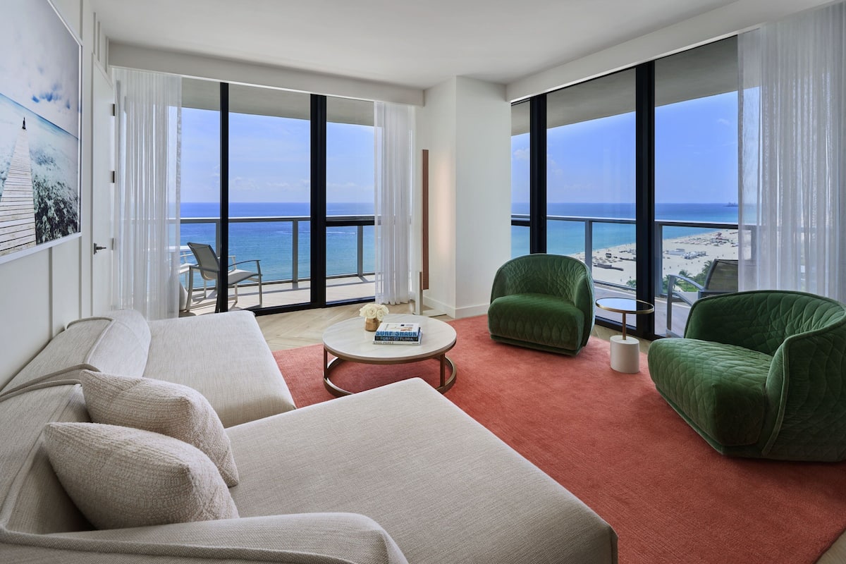 A spacious living room with a large window overlooking a beach, a green armchair, a beige sofa, and a small coffee table.
