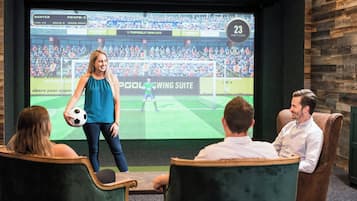 A person standing in front of a large screen displaying a soccer game, with three seated individuals watching.