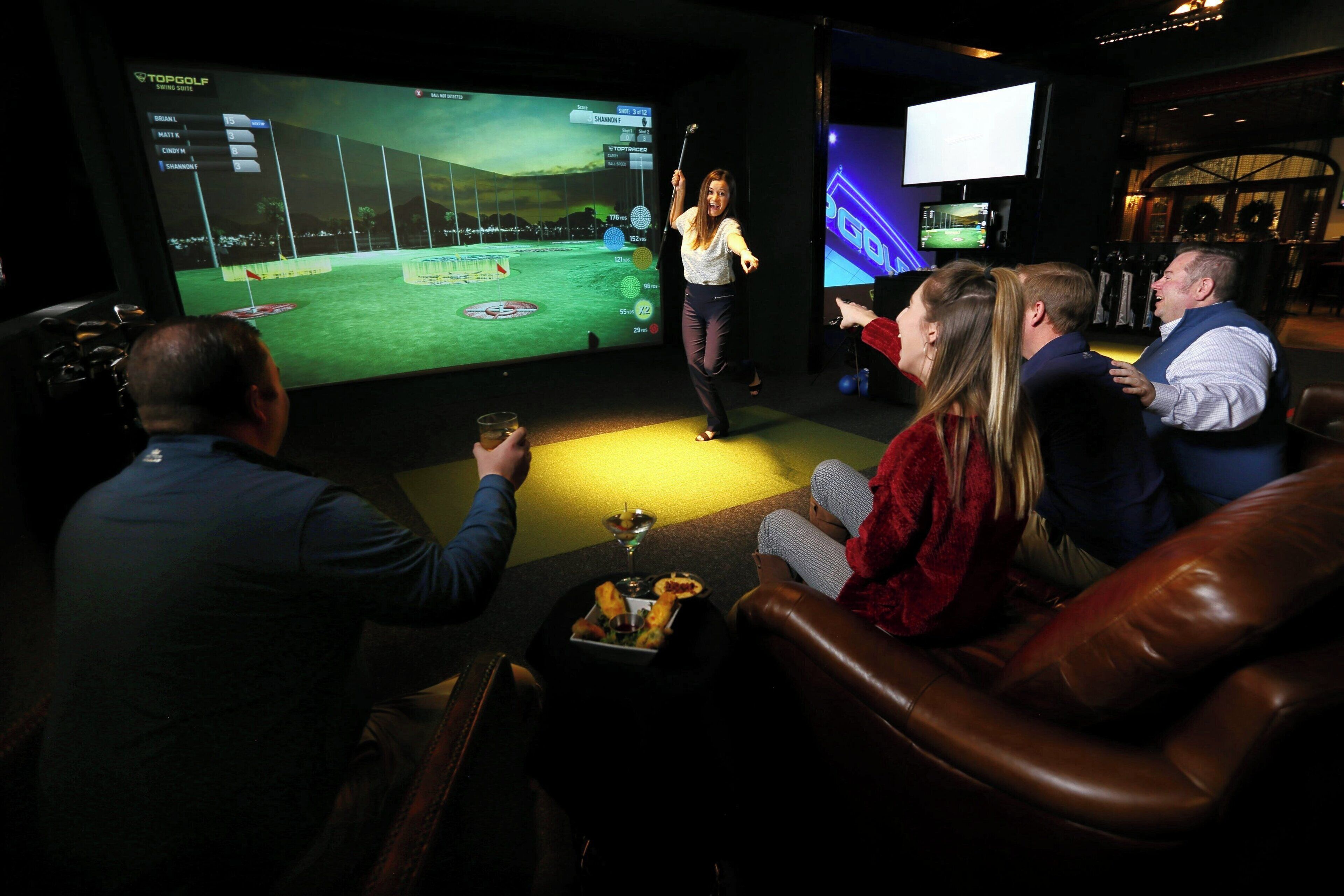 Eine Gruppe von Personen beobachtet ein Golf-Simulationsspiel auf einem großen Bildschirm in einem schwach beleuchteten Raum mit Ledersitzmöbeln.