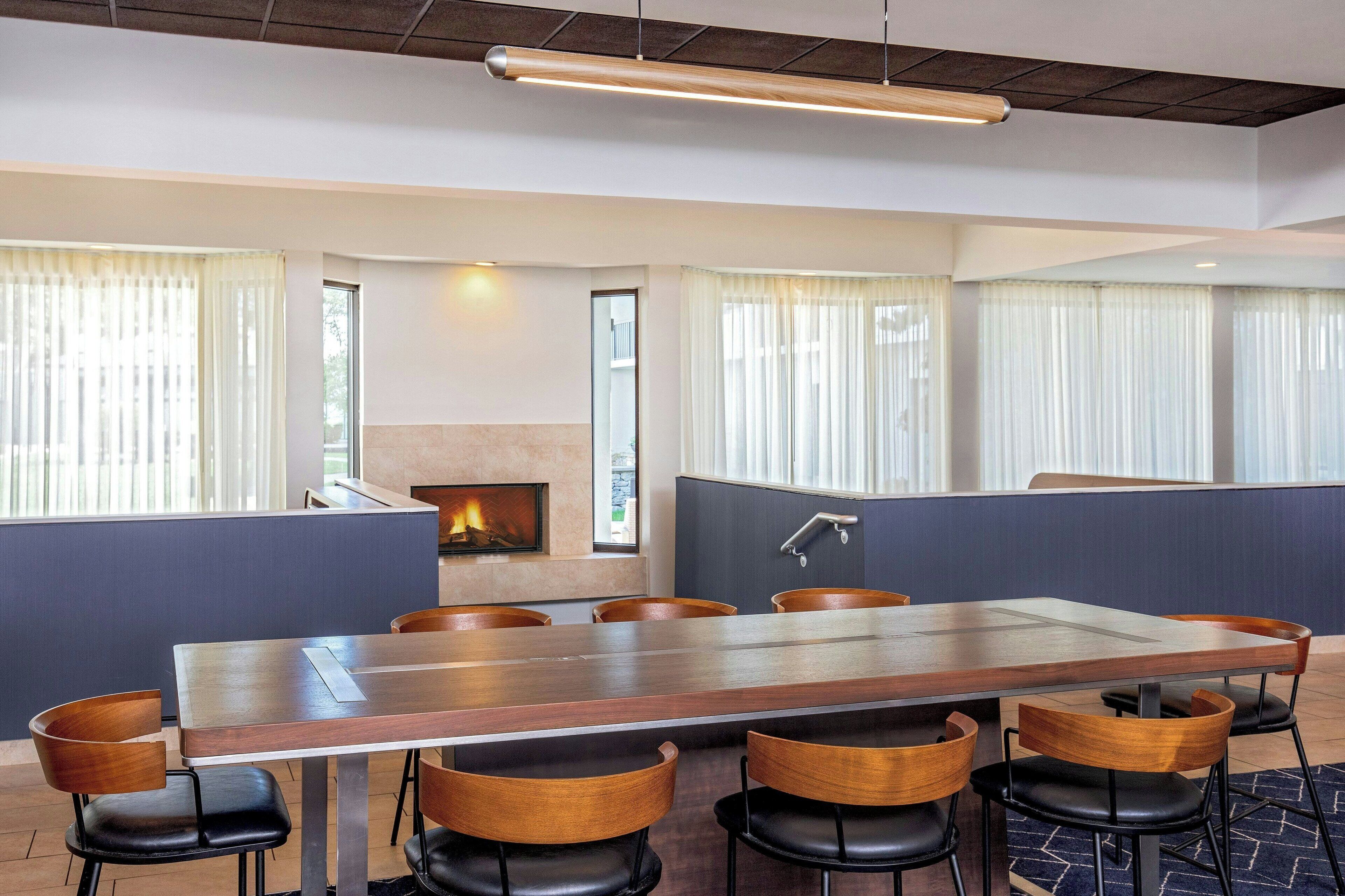 A conference room with a long table, chairs, a fireplace, and large windows with curtains.