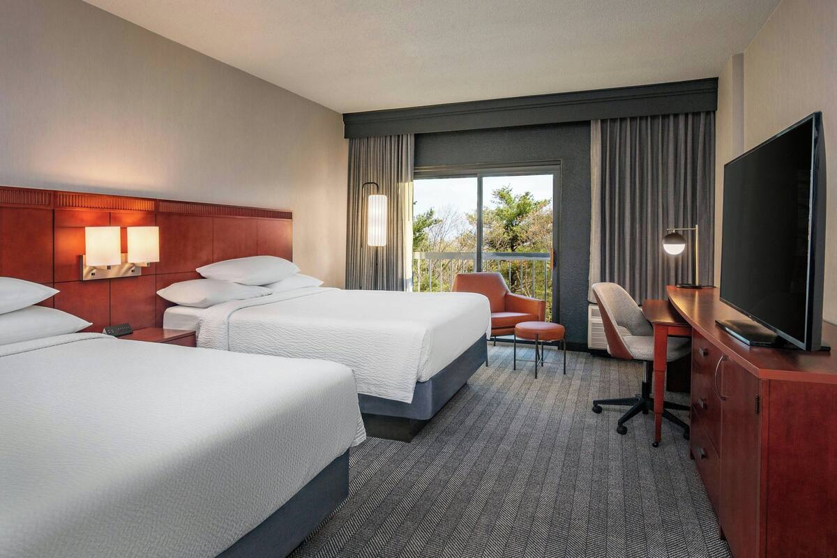 A hotel room with two beds, a desk, a chair, a TV, and a balcony with curtains.