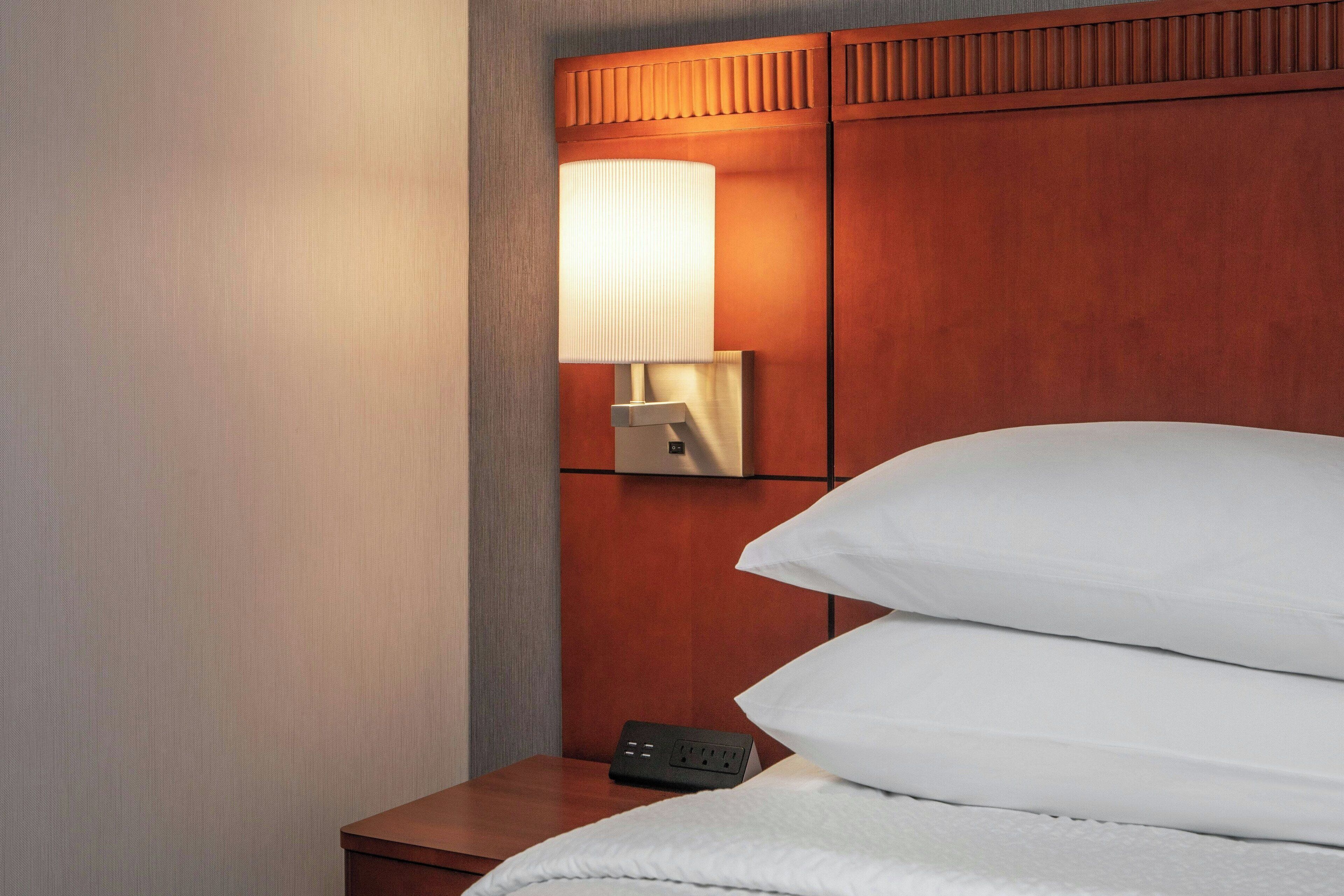 A neatly made bed with white linens, a bedside table with a lamp, and a power outlet.