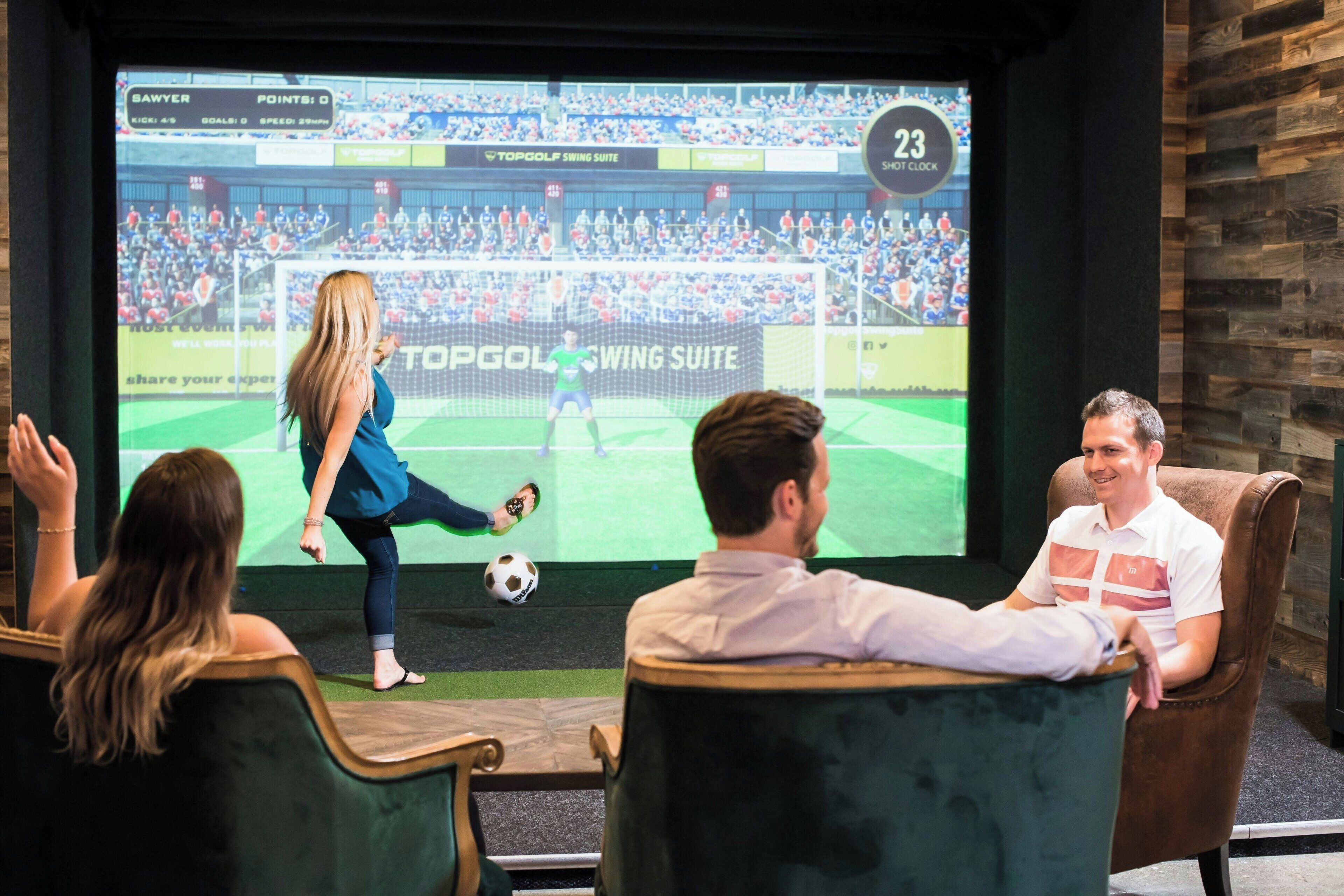 A room with a large screen displaying a soccer game, a person playing soccer, and three seated individuals watching.