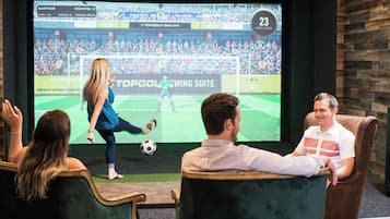 A room with a large screen displaying a soccer game, a person playing soccer, and three seated individuals watching.
