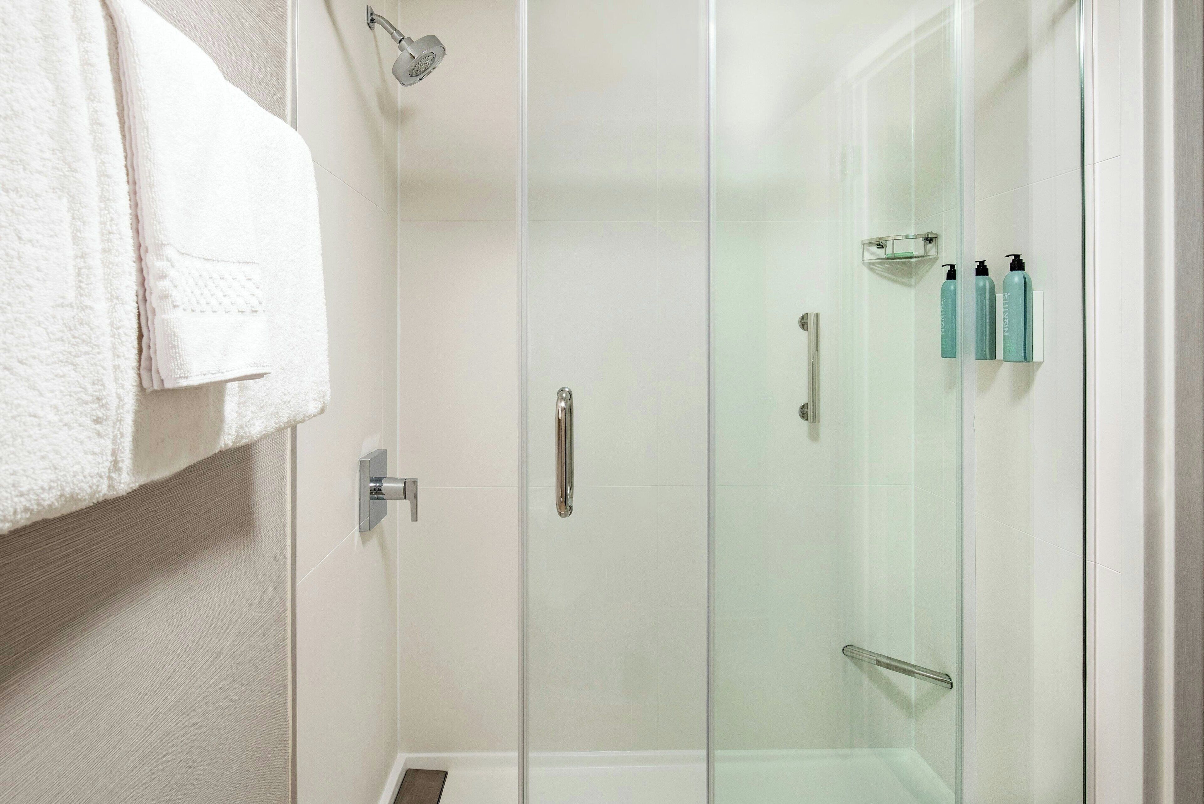A modern bathroom with a glass shower enclosure, a white towel hanging on the wall, and toiletries on a shelf.