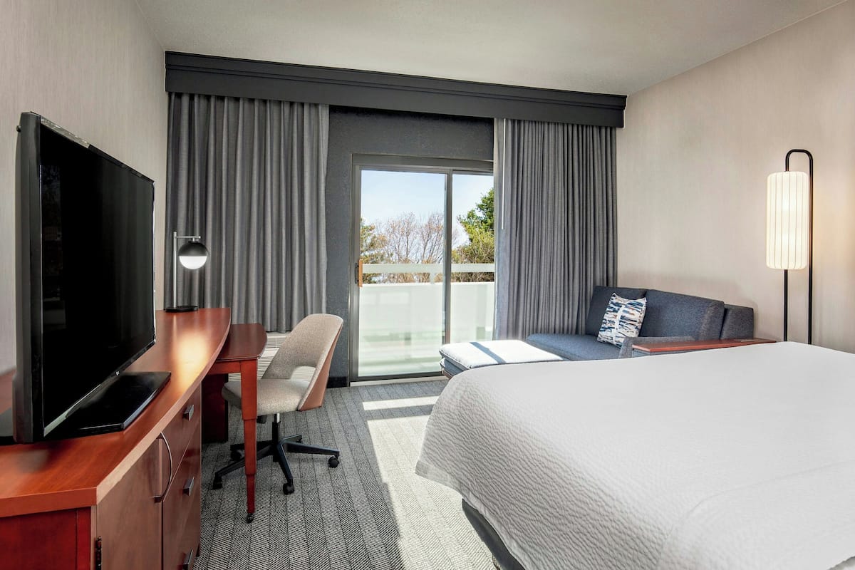 A hotel room with a bed, a desk with a TV, a chair, and a balcony with curtains.