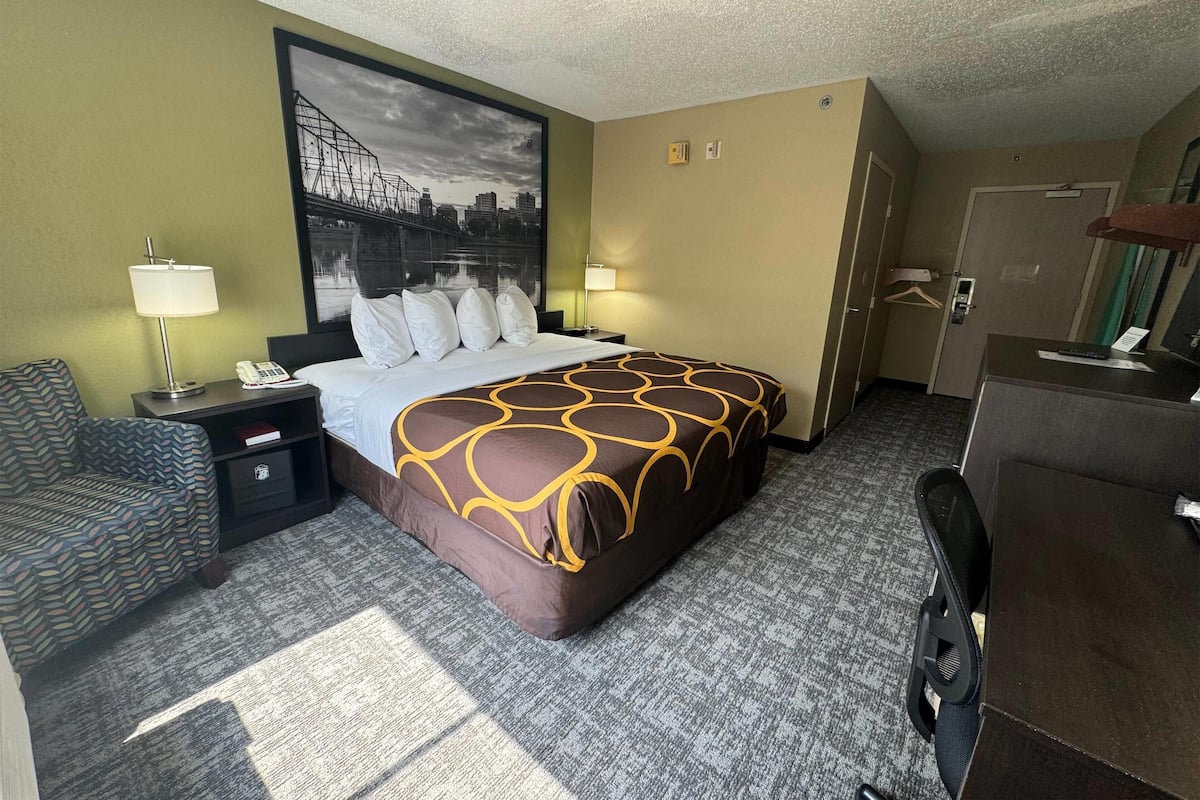 Une chambre d’hôtel avec un lit, deux tables de chevet, une chaise, un bureau et un grand tableau représentant un paysage urbain accroché au mur.