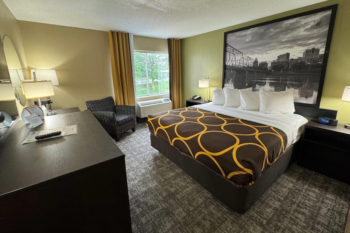 Une chambre d’hôtel avec un grand lit, un bureau, une chaise, une fenêtre avec des rideaux et un tableau représentant un paysage urbain accroché au mur.